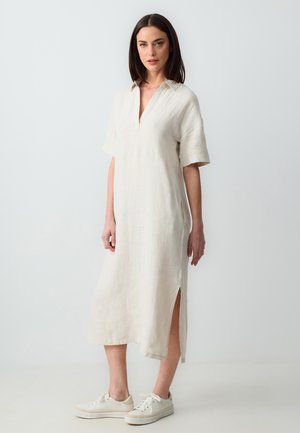 Beiges Leinenkleid mit V-Ausschnitt, kurzen Ärmeln, seitlichen Schlitzen und lockerem Schnitt, kombiniert mit weißen Sneakers. Glatte Textur und leicht.