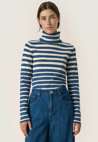 Plavi i kremasti prugasti turtleneck, rebraste teksture, dugih rukava, u kombinaciji s trapericama širokih nogavica koje imaju prednje džepove i zatvaranje na dugme.