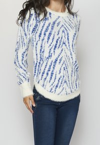 Fluffy white sweater with blue zigzag pattern, round neckline, and ribbed cuffs, paired with dark blue jeans featuring an embellished design.