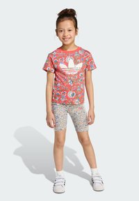 Röd blommönstrad t-shirt med vit logotyp, tillsammans med flerfärgade, mönstrade cykelshorts. Modellen bär vita löparskor.