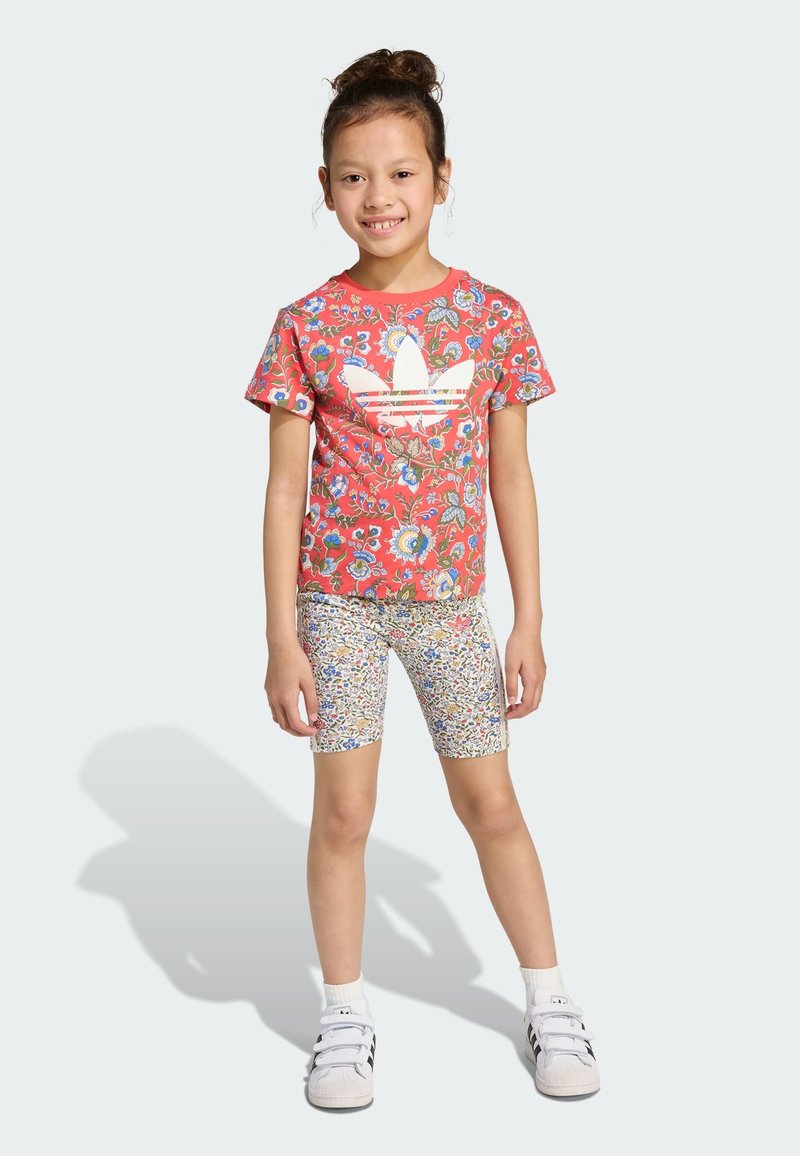 Röd blommönstrad t-shirt med vit logotyp, tillsammans med flerfärgade, mönstrade cykelshorts. Modellen bär vita löparskor.