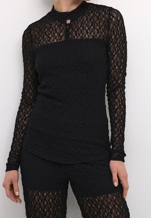 Kvinde iført tætsiddende sort bluse i blonder med lange ærmer og matchende blondeshorts mod ensfarvet hvid baggrund.