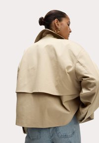 Beige cropped jas met een hoge kraag, gelaagd ontwerp aan de achterkant en een zachte textuur. Gecombineerd met lichtblauwe high-waisted jeans.