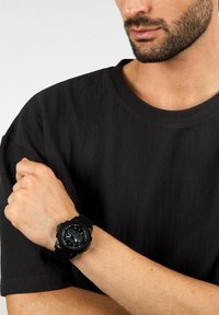 Orologio nero con quadrante rotondo, display digitale, cinturino in gomma e marcature audaci. Indossato su un polso contro uno sfondo di tessuto scuro.