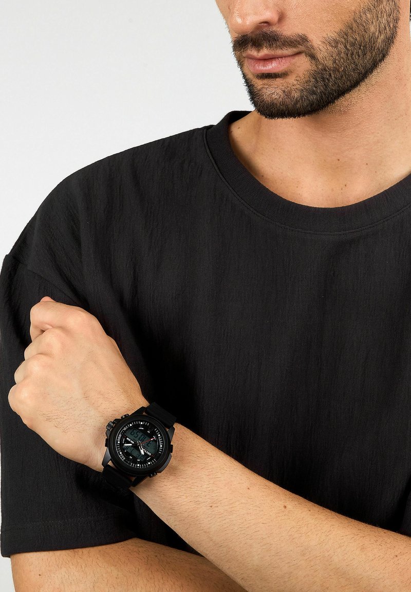 Orologio nero con quadrante rotondo, display digitale, cinturino in gomma e marcature audaci. Indossato su un polso contro uno sfondo di tessuto scuro.