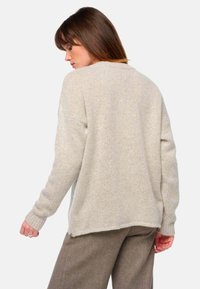 Beige übergroßer Pullover mit strukturiertem Strick, gerippten Bündchen und einem etwas längeren hinteren Saum. Kombiniert mit gemusterten braunen Hosen.