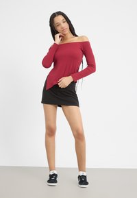Off-shoulder burgunderfarget topp med lange ermer, ribbestruktur, asymmetrisk kant; kombinert med et svart miniskjørt og svarte sneakers.