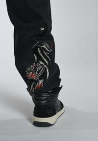 Los pantalones de mezclilla negros cuentan con un gran diseño bordado de un pantera en gris y rojo en el costado. El tobillo revela una zapatilla de caña alta en blanco y negro.