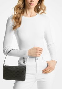 Černá kabelka s monogramem, elegantního obdélníkového tvaru, vyrobená z kůže, s krátkým popruhem, nošena k bílému topu s dlouhým rukávem a džínám.