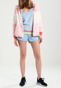 Giacca sportiva rosa, bianca e azzurro chiaro con cerniera, con polsini rossi a contrasto, abbinata a una canotta azzurra chiara, pantaloni shorts in denim e sneaker nere.