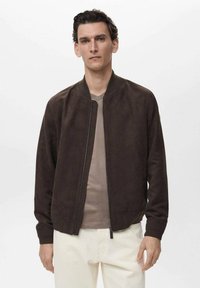 Braune Wildleder Bomberjacke mit Reißverschluss, gerippten Bündchen und Kragen. Kombiniert mit einem hellbeigen Hemd und off-white Hosen.
