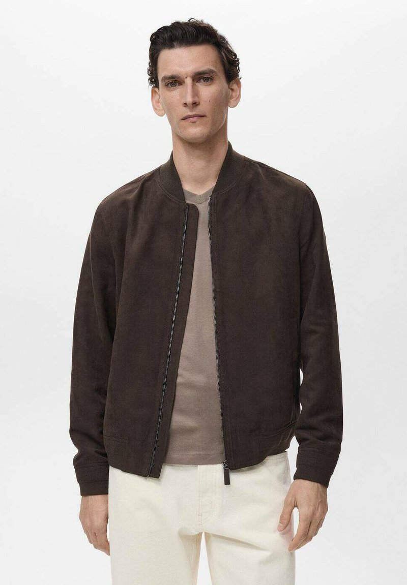 Braune Wildleder Bomberjacke mit Reißverschluss, gerippten Bündchen und Kragen. Kombiniert mit einem hellbeigen Hemd und off-white Hosen.