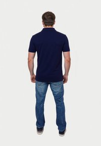 Young man facing away, wearing a navy polo and blue jeans, standing against a plain white background. Casual attire, relaxed posture.