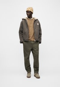 Άνδρας που φοράει μπεζ σκουφάκι με στίγματα, καφέ τζάκετ με επένδυση φλις, μπεζ φούτερ, παντελόνι ελιάς και μπεζ μπότες, στέκεται με τα χέρια στις τσέπες.