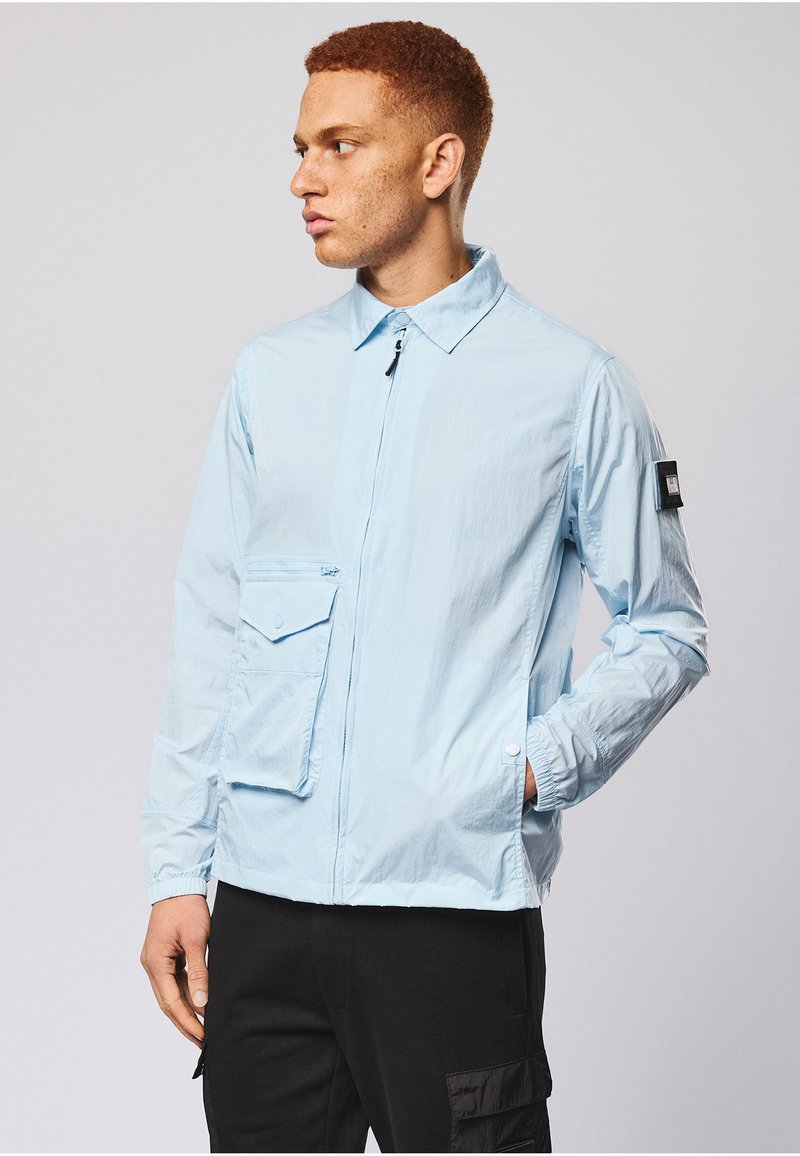 Jeune homme aux cheveux roux portant une veste zippée bleu clair avec plusieurs poches et un pantalon cargo noir, regardant vers la gauche.