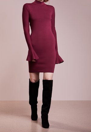 Femme portant une robe bordeaux ajustée à manches longues avec des poignets évasés et des bottes noires hautes jusqu'aux genoux, debout devant un fond neutre.