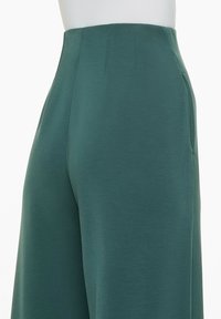 Pantalons larges verts en tissu lisse. Présentent une taille haute avec des poches latérales et une texture subtile. Aucun fermoir visible.