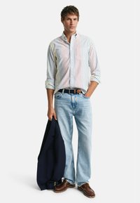 Gestreifte Hemd mit Knöpfen in Pastellfarben, hellblaue Jeans, dunkler Blazer über dem Arm, braune Schnürschuhe. Lässiges Outfit.