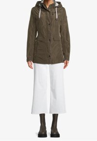 Lekka kurtka w oliwkowej zieleni z kapturem, przednimi kieszeniami i zamkami błyskawicznymi, zestawiona z białymi szerokimi spodniami i ciemnymi wodoodpornymi butami.