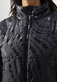 Black puffer jacket with shiny, embossed "HUGO" logo pattern. Features a stand-up collar and front zipper. Smooth texture, lightweight design.