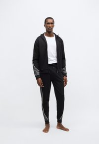 Čierna športová súprava obsahujúca mikinu s kapucňou na zips so šedými akcentmi na rukávoch a zodpovedajúce nohavice so sťahovacou šnúrkou a bielymi pruhmi na bokoch.