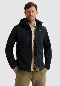 Mann trägt schwarzen Reißverschlussjacke über beige Hemd mit Knöpfen und Khakihose, steht mit Händen in den Taschen vor einfachem Hintergrund.