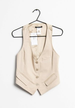 Gilet beige sans manches à boutons sur le devant, avec trois boutons, deux poches à rabat et une poche poitrine, suspendu à un cintre en fil noir.