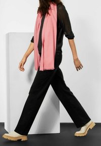 Zwarte jeans en een zwarte top gecombineerd met een lichtroze sjaal. Kenmerkt zich door beige chunky schoenen met een ribbelige zool, geplaatst tegen een minimalistische achtergrond.