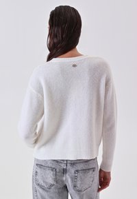 Maglione bianco a maglia testurizzata con una vestibilità leggermente accorciata e orlo a coste. Presenta un piccolo dettaglio del logo vicino al collo sul retro.