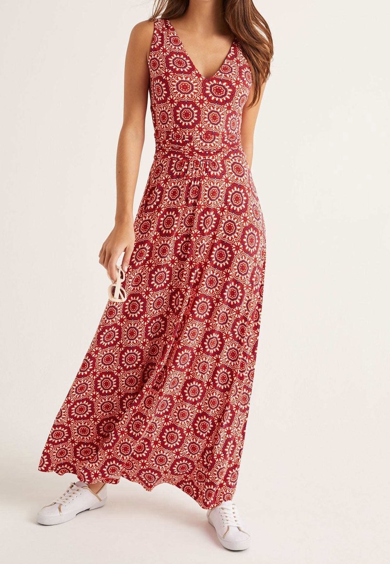 Femme portant une robe maxi rouge sans manches avec des motifs floraux circulaires blancs, tenant des lunettes de soleil, assortie à des baskets blanches.