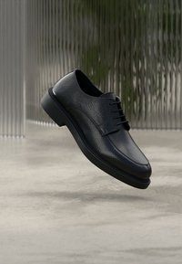 Schwarzer Lederschuh mit strukturierter Oberfläche, abgerundeter Spitze und Schnürdesign. Verfügt über eine dicke Sohle für zusätzlichen Komfort und Unterstützung.