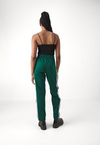 Vihreät urheiluhousut, joissa on valkoiset sivuraidat, joustava vyötärö ja sileä pinta. Yhdistetty mustan tiukasti istuvan paidan ja mustien jalkineiden kanssa.