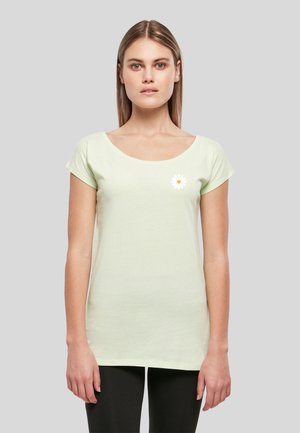 Jeune femme aux cheveux lisses porte un t-shirt vert clair à manches courtes avec un petit motif marguerite blanche sur la poitrine gauche, pantalon noir.