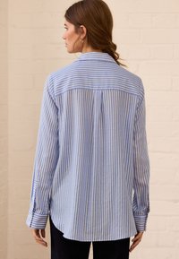 Mėlynos ir baltos spalvų dryžuota ilgomis rankovėmis marškiniai su tiesiu apdailos kraštu, užsegamais rankogaliais ir atlošo skydeliu nugaroje. Lygios tekstūros audinys.