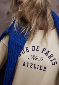 Beżowa bluza z napisem "Avenue de Paris No. 9 Atelier", zestawiona z teksturowanym, niebieskim dzianym szalikiem, prezentująca casualowy styl warstwowy.