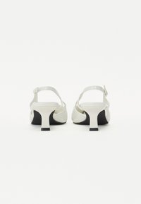 Sandales blanches à talons hauts avec une sangle arrière, présentant une finition brillante, une forme incurvée et un talon noir contrasté. Détail de boucle simple.