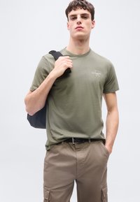 Νεαρός άνδρας που φοράει ελιάς πράσινη μπλούζα Calvin Klein και καφέ παντελόνια cargo, κρατώντας με το ένα χέρι το λουρί μιας μαύρης τσάντας πλάτης.