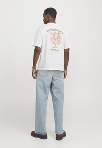 Witte katoenen T-shirt met een palmboomgrafiek in het oranje op de achterkant. Draagt het met lichtblauwe, ruimvallende jeans en bruine schoenen.
