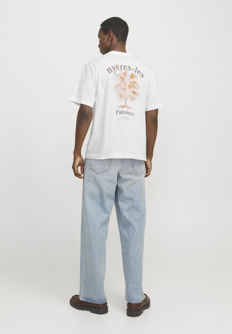 Camiseta blanca de algodón con un gráfico de palmera en color naranja en la parte trasera. Se lleva con jeans holgados de color azul claro y zapatos marrones.