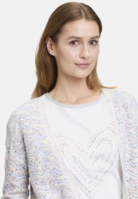 Flerfärgad virkad cardigan med ett strukturerat vågmönster, bärs över en vit T-shirt med ett silverhjärtemönster och rhinstenar.