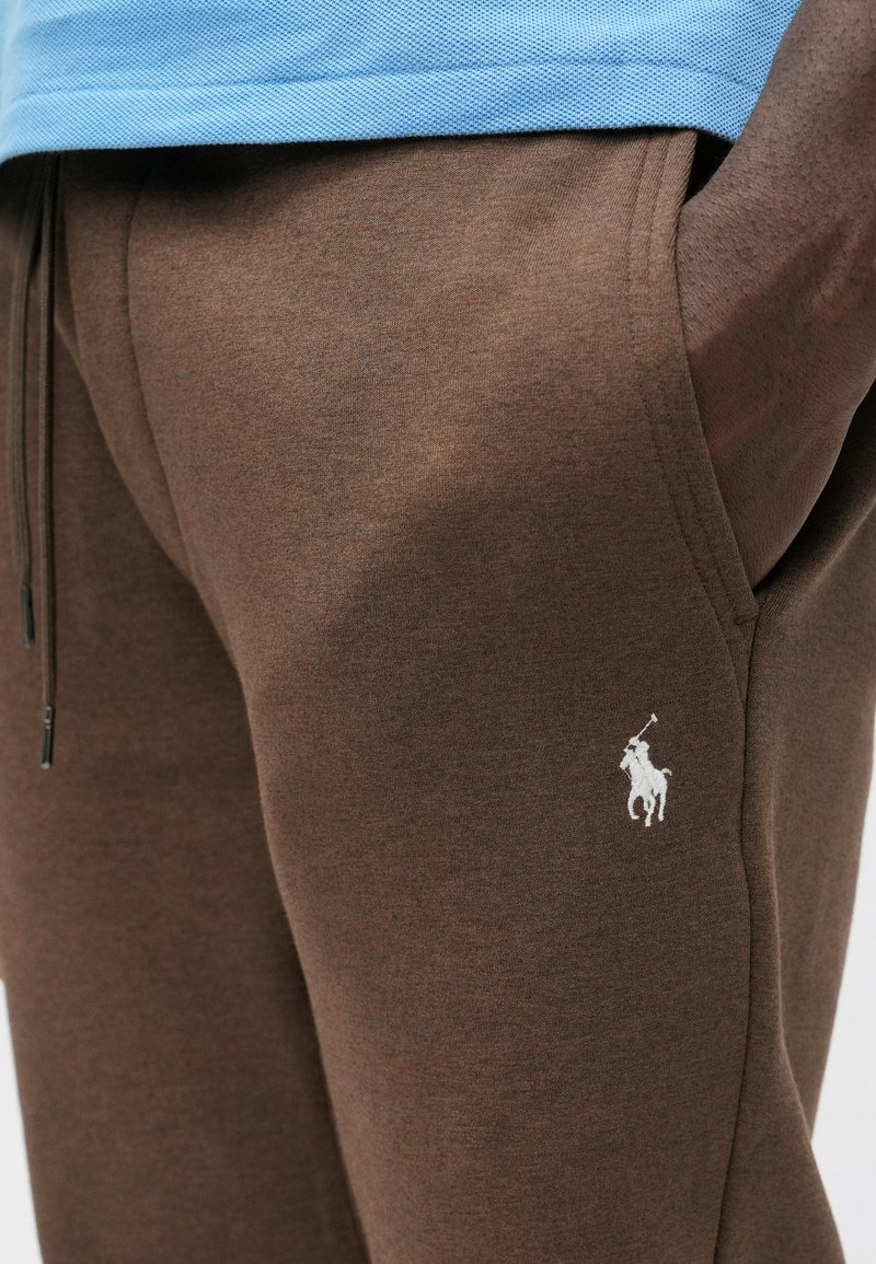 Braune Jogginghose aus weichem Material, mit einem Kordelzugbund und einem weißen Logoemblem auf dem Oberschenkel. Die Textur wirkt glatt.