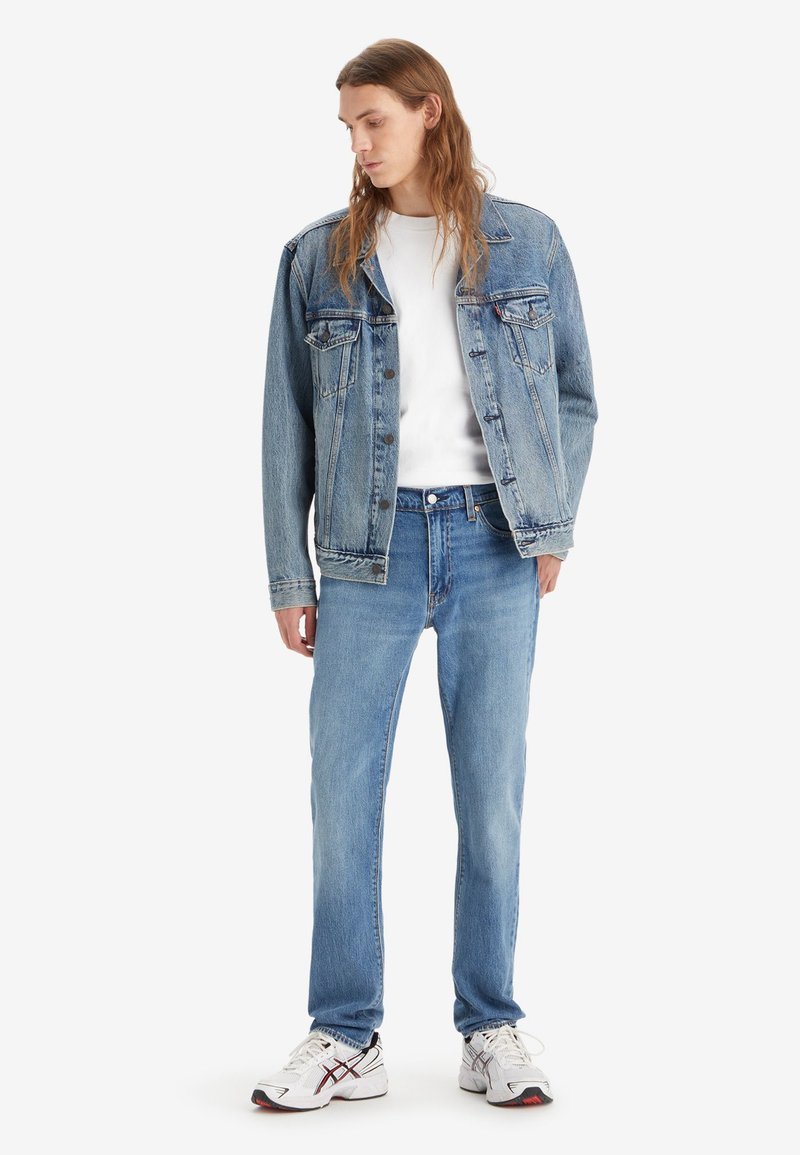 Chaqueta de mezclilla azul con cierre de botones, combinada con una camisa blanca y jeans de mezclilla azul claro. Zapatillas blancas con acentos en negro y rojo.