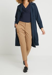 Lång marinblå kappa över en mörk överdel, matchad med ljusbruna skräddarsydda byxor. Svarta loafers fulländar outfiten. Släta texturer, klassisk passform.