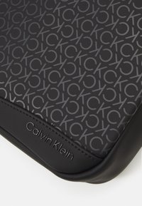 Accesorio negro de Calvin Klein con un sutil patrón texturizado de letras entrelazadas. Bordes suaves con detalle de logo en relieve.