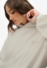 Felpa crema realizzata in tessuto a costine, con cappuccio regolabile e un logo ricamato discreto sul petto. Morbida al tatto e dal fit rilassato.