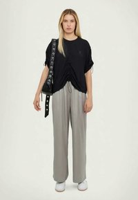 Femme aux longs cheveux blonds portant un haut noir froncé, un pantalon gris ample, des baskets blanches et un sac à bandoulière noir.