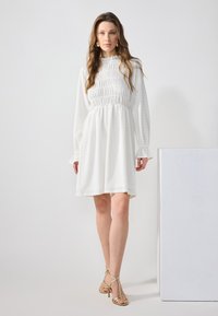 Bílé šaty s dlouhými rukávy, řaseným živůtkem a sukní ve tvaru A. Woven texturou, mírným leskem, spárované se zlatými sandály na vysokém podpatku.
