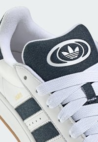 Adidas Campus sneakers au o bază din piele albă, o parte superioară din denim închis, șireturi albe și un accent cu logo rotund pe limbă. Textul 'CAMPUS' este aurie.
