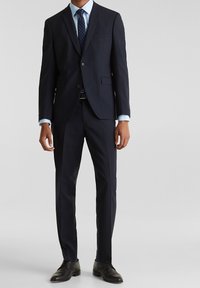 Marinblå kostym med fast struktur, enkelknäppt kavaj, slagkrage, byxor med rak fram, kombinerad med en ljusblå skjorta och prickig slips.