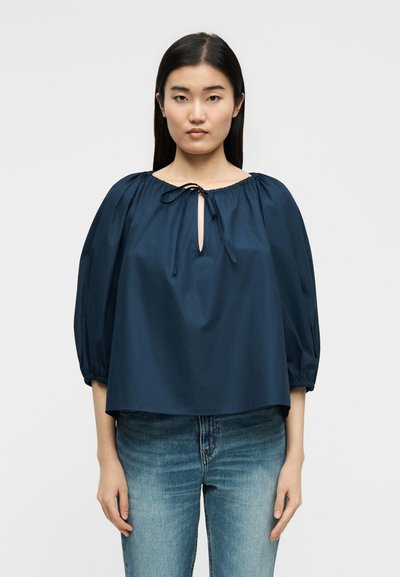 Mladá žena s dlhými tmavými vlasmi, oblečená v voľnej námorníckej blúzke s 3/4 nafúknutými rukávmi a svetlomodrých džínsoch, stojaca pred jednoliatym pozadím.